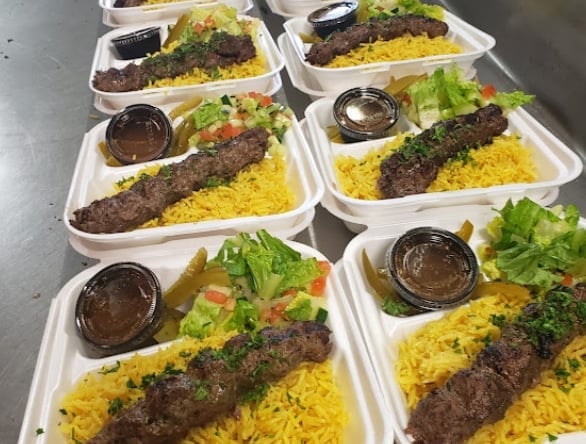 Six takeout containers with grilled meat kebab skewers, yellow rice, fresh lettuce, and sauce cups arranged on a table