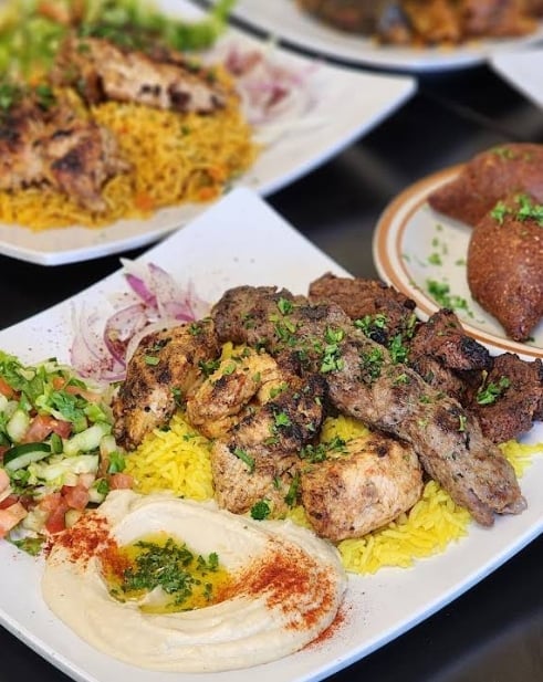 Grilled lamb kebabs with yellow rice, hummus, fresh salad, and herbs on white plate in Middle Eastern restaurant